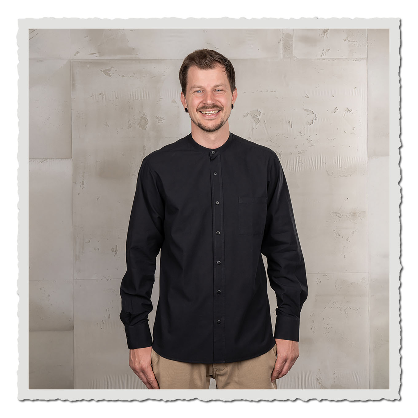 Person mit dunklem Hemd und heller Hose vor hellem, neutralem Hintergrund im Studio.
